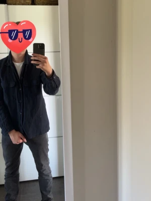 Svart overshirt med fickor - Stilren svart overshirt med knäppning framtill och två bröstfickor. Skjortan har långa ärmar med manschett och är tillverkad i ett linne tyg som ger en avslappnad look. Perfekt att bära över en t-shirt eller hoodie för en casual stil.