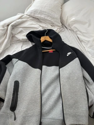 Nike Tech Fleece hoodie svart/grå - Nike Tech Fleece hoodie i svart och grått med dragkedja och huva. Snygg tvåfärgad design, vit Nike-logga på bröstet och praktisk ficka med dragkedja på ena ärmen. Perfekt för en sportig och avslappnad stil.