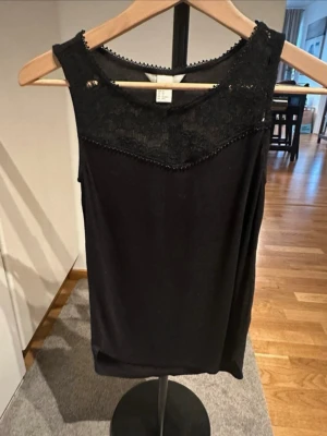 Svart linnetopp med spetsdetalj - Snygg svart ärmlös linnetopp med spets upptill och diskreta pärldetaljer längs halsringningen. Toppen har en rak passform och är perfekt att styla med jeans eller kjol. Materialet känns mjukt och följsamt mot huden.