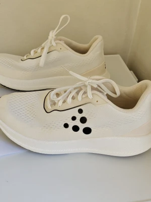 Vita sneakers från Craft - Stilrena vita sneakers från Craft med svart logotyp på sidan. Ovandelen är i meshmaterial för bra andningsförmåga och skon har en tjock, vit sula för extra komfort. Klassisk snörning och låg modell som passar till många olika outfits. Modellen heter Pacer i 44 storlek.  Välskötta och sparsamt använda .