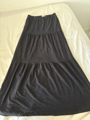 Svart långkjol med broderade detaljer - Snygg svart långkjol från Kappahl med broderade hålmönster och resår i midjan. Kjolen har flera volanger och är tillverkad i ett mjukt tyg som känns skönt mot huden.