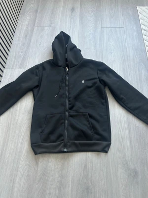 Svart hoodie från Polo Ralph Lauren - Svart hoodie med dragkedja från Polo Ralph Lauren. Klassisk design med huva och dragsko, två fickor framtill och den ikoniska vita loggan broderad på bröstet. Perfekt för en avslappnad och stilren look.Tröjan är helt ny och oanvänd! storleken är M