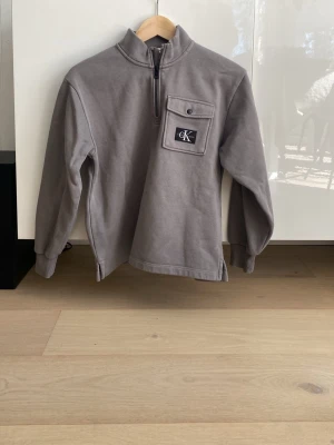 Grå Calvin Klein tröja med halv dragkedja - Snygg grå långärmad tröja från Calvin Klein med halv dragkedja och hög krage. Bröstficka med lock och CK-logga framtill. Tröjan har ribbade muddar vid ärmslut och är tillverkad i mjukt material.