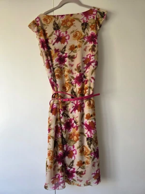 Esprit blommig klänning med nätt skärp - Långklänning med blommigt mönster i rosa, orange och grönt på ljus botten. Klänningen har en smal rosa skärp i midjan och är ärmlös med rund halsringning. Perfekt för sommarens varma dagar.