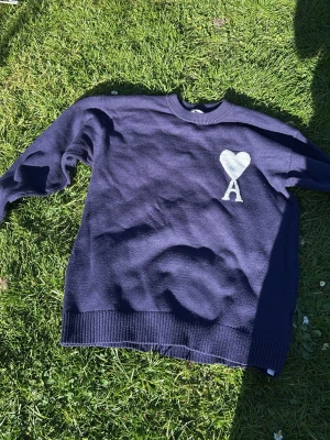 Mörkblå stickad tröja från Ami Paris - Mörkblå stickad tröja från Ami Paris med vitt hjärta och bokstaven A broderat på bröstet. Tröjan har rund halsringning, långa ärmar och ribbade muddar vid ärmslut och nederkant. Perfekt för en stilren och trendig look.