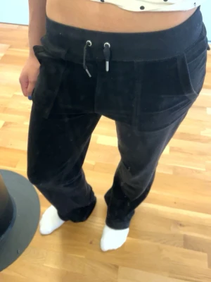 Svarta juciy couture byxor - Svarta mjukisbyxor i mjuk velour med dragsko i midjan och stora fickor framtill. Byxorna har en avslappnad passform och raka ben, perfekta för en chill och bekväm stil. Byxorna har kläd tejp innanför för att dem var långa för mig förut o då korta jag ner dem me klädtejp. Orginal pris 1000kr