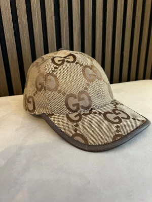 Beige keps från GGmed GG-mönster - Snygg beige keps från Gucci med klassiskt GG-monogram i brunt över hela kepsen. Justerbar rem baktill i brunt läder och böjd skärm. Tillverkad i canvasmaterial med exklusiv känsla. Perfekt accessoar för en trendig look.
