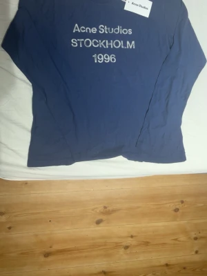 Blå långärmad topp Acne Studios - Säljer en blå långärmad topp från Acne Studios med tryck 'STOCKHOLM 1996' framtill. Enkel och clean design, perfekt för dig som gillar stilrena plagg med en touch av streetwear. Skön passform och mjukt material.