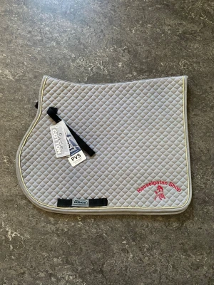 Grått schabrak från Equest - Säljer ett grått schabrak från Equest, modell PVS för ponny. Schabraket är tillverkat i 100% bomull med quiltat rutmönster och har ljus kant samt broderad text 'Hasselgatan Shop' i rött. Perfekt för allroundridning.