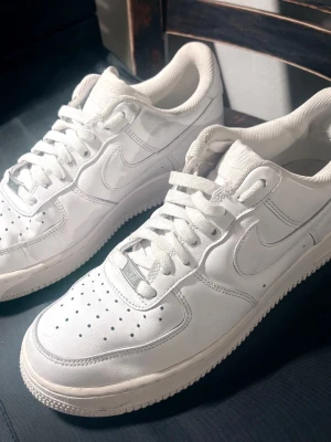 Nike Air Force 1 vita sneakers - Klassiska helvita Nike Air Force 1 sneakers i läder med perforerad tå, vit sula och ikonisk Swoosh-logga på sidorna. Skorna har lågt skaft, platt snörning och diskret Air-detalj på sulan. Perfekta för en clean och stilren look.