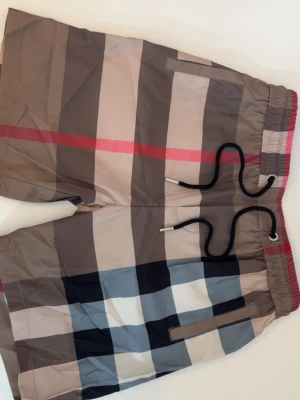 Rutig shorts med snörning - Snygga rutiga shorts från Burberry med klassiskt mönster i beige, svart, vitt och rött. Shortsen har elastisk midja med svart snörning och praktiska fickor. Perfekta för en trendig och avslappnad stil.