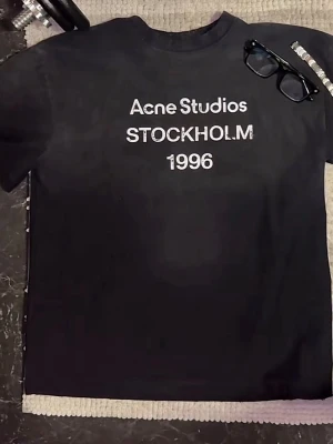 Svart Acne Studios t-shirt - Säljer en svart t-shirt från Acne Studios med vit text på bröstet där det står 'Acne Studios STOCKHOLM 1996'. Klassisk rund hals och korta ärmar. Perfekt till jeans eller shorts för en clean och stilren look.