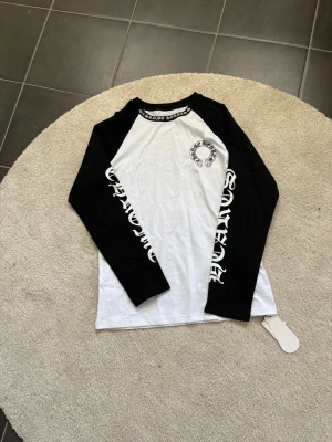 Chrome Hearts långärmad topp - Snygg långärmad topp från Chrome Hearts med svartvita kontraster. Svarta ärmar med vit text längs båda armarna och logga på bröstet. Klassisk baseball-modell med rund halsringning och tryckdetaljer. Perfekt för en streetwear-look.