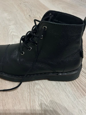 Svarta Levis kängor i skinn med snörning - Snygga svarta kängor i äkta skinn från Levis med grov sula och klassisk snörning framtill. Kängorna har rund tå, metallöljetter och en dragflik bak för enkel påtagning. Perfekta för en edgy och stilren look.