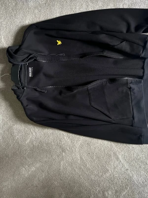 Svart hoodie från Lyle & Scott - Svart hoodie med dragkedja från Lyle & Scott. Klassisk design med huva, känguruficka och gul logga på bröstet. Perfekt för en chill och stilren look. Tillverkad i mjukt material som känns skönt mot huden.