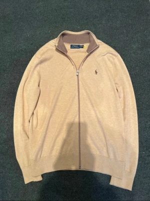 Beige ziptröja från Polo Ralph Lauren - Snygg beige ziptröja från Polo Ralph Lauren med krage och diskret logga på bröstet. Tröjan har hel dragkedja och ribbade muddar vid ärmslut och nederkant. Perfekt för lager-på-lager och enkel att matcha med det mesta. Ovh. 220€