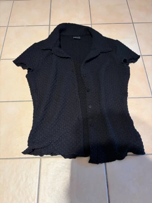Svart kortärmad blus med krage - Snygg svart blus med korta ärmar och klassisk krage. Blusen har knäppning framtill och är gjord i ett stretchigt, krinklat tyg som ger en cool struktur. Perfekt att styla till jeans eller kjol för en trendig look.