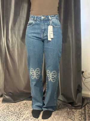 Blå raka jeans med broderi från Gina Tricot - Snygga blå jeans med raka ben och hög midja från Gina Tricot. Jeansen har vitt broderi i form av fjärilar på båda benen framtill och klassiska bakfickor. Oanvänd 
