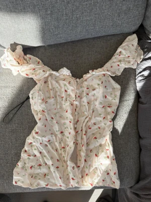 Söt blommig blus med volangärmar - Romantisk blus i ljus färg med små blommor och volangiga offshoulder-ärmar. Toppen har knappar framtill och en lätt rynkad struktur som ger en feminin känsla. Perfekt för dig som gillar en gullig och trendig stil.