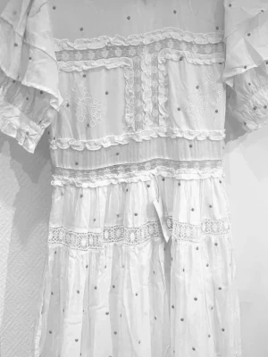 By malina maxiklänning - Romantisk vit långklänning med broderade spetsdetaljer, volanger och små prickar. Klänningen har korta ärmar, vackra spetsband och ett luftigt, flowy fall. Perfekt för sommarens alla tillfällen och ger en bohemisk känsla. Storlek medium, nyskick med lappar 