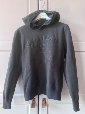 Grå hoodie från Zadig & Voltaire - Snygg grå hoodie från Zadig & Voltaire med tryck på bröstet där det står 'Rive Droite', 'Art is Truth' och 'Est. 1997'. Hoodien har en klassisk huva och ribbade muddar. Perfekt för en avslappnad och cool stil.
