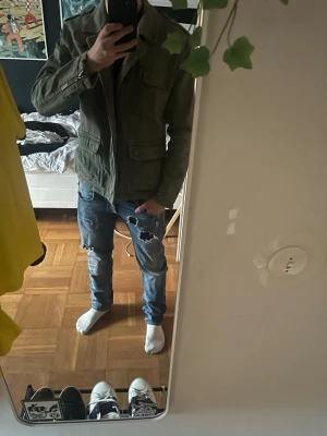 Olivgrön overshirt med fickor - Fin overshirt med flera fickor och dragkedjor! Snygg sköld på bröstet och regular i storleken. Liten defekt på armen (se bild). Jag är 176 cm  63 kg. 