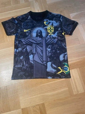 Nike Brasil matchtröja svart/gul - Säljer en svart matchtröja från Nike med Brasilien-tema. Tröjan har ett stort tryck av Kristusstatyn, brasilianska flaggan och detaljer i gult. Korta ärmar och rund hals. Perfekt för dig som älskar fotboll och vill sticka ut på planen.