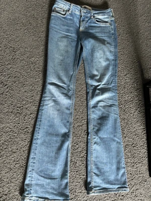 Ljusblå bootcut jeans - Säljer ett par ljusblå bootcut jeans med klassisk femficksdesign och låg midja. Jeansen har utsvängda ben och är tillverkade i denim med snygga slitningar framtill. Perfekta för dig som gillar en retro och avslappnad stil.