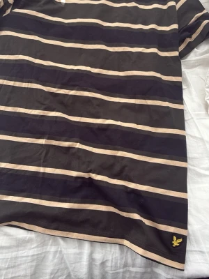 Randig t-shirt från Lyle & Scott - Snygg svart t-shirt med beigea ränder från Lyle & Scott. Klassisk passform och korta ärmar. Ikonisk gul logga broderad längst ner. Perfekt till jeans eller shorts för en chill look.
