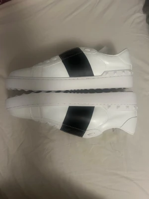 Valentino Garavani vita sneakers 44 - Säljer ett par vita sneakers från Valentino Garavani med bred svart rem över vristen och ikoniska nitar längs hälen. Skorna är i slätt läder och har en chunky vit sula. Perfekta för dig som gillar stilrena men ändå unika detaljer. i 44 SKRIV PÅ 😎‼️
