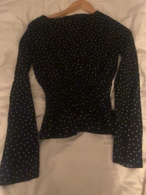 Svart prickig blus med smock - Trendig svart blus med vita småprickar och smockad midja som ger en snygg siluett. Blusen har långa ärmar och lätt utsvängd nederkant. Perfekt att styla med jeans eller kjol för en cool look.