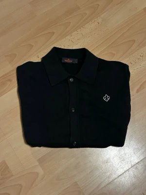 Kortärmad Morris skjorta - Storlek M passar S också .Klassisk mörkgrå skjorta från Morris med knappar framtill och vit broderad logga på bröstet. Skjortan har en stilren krage och är perfekt för dig som gillar enkel och tidlös stil.