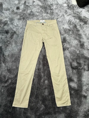 Beige chinos från Hampton Republic - Snygga beige chinos från Hampton Republic med klassisk rak passform. Byxorna har framfickor, bälteshällor och knäppning med knapp och dragkedja. Tillverkade i mjuk bomull för en bekväm känsla. Perfekta för en stilren och avslappnad look. Storlek 158.