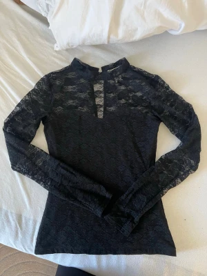 Svart spetsblus med lång ärm - Snygg svart blus i spets med lång ärm och hög krage. Blusen har en liten v-ringning framtill och är helt genombruten i ett blommigt spetsmönster. Perfekt att styla med jeans eller kjol för en ute kväll me tjejerna.💕