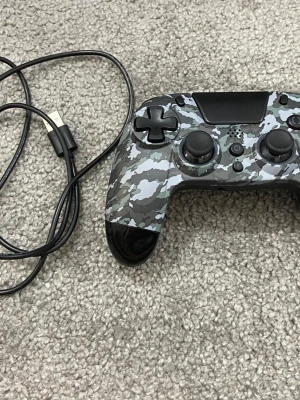 Kamouflage handkontroll med USB-kabel - Snygg handkontroll med grått kamouflagemönster och svarta detaljer. Ergonomisk design med två analoga spakar, styrkors och flera knappar. Kommer med USB-kabel för laddning eller anslutning. Perfekt för gaming på konsol eller PC.