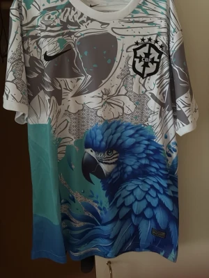 Nike Brasil Dri-Fit t-shirt med blå papegoja - Säljer en unik Nike Brasil Dri-Fit t-shirt med stort tryck av en blå papegoja och tropiska blommor. Tröjan är vit, grå och blå med CBF-logga och Nike Swoosh på bröstet. Perfekt för dig som gillar färgstarka sportkläder och snygg design.