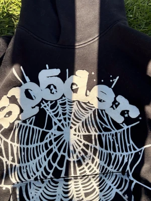 Svart hoodie med spindelnäts-tryck - Svart hoodie från Sp5der med stort vitt spindelnäts-tryck och graffiti-inspirerad logga på ryggen. Hoodien har huva och tryck med stjärnor på ena ärmen. Perfekt för dig som gillar streetwear och unika prints.