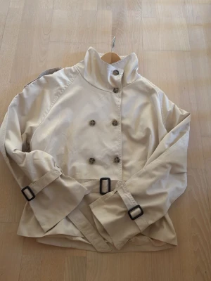 Beige kort trenchcoat med bälten - Stilren beige trenchcoat med hög krage, dubbelknäppning och svarta spännen vid ärmsluten. Den är helt ny och har aldrig använt den då den är för stor på mig.