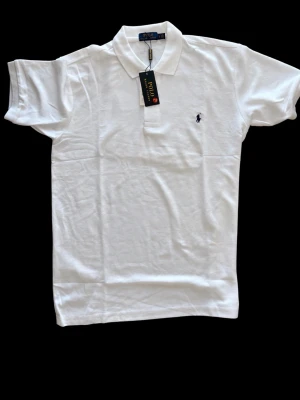 Vit pikétröja från Polo Ralph Lauren - Klassisk vit pikétröja från Polo Ralph Lauren med krage och korta ärmar. Tröjan har den ikoniska broderade loggan på bröstet och knäppning vid halsen. Perfekt för en stilren och tidlös look.