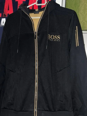 Svart hoodie från Hugo Boss - Svart hoodie från Hugo Boss med dragkedja och huva. Guldiga detaljer på dragkedjan och loggan på bröstet samt dekorativa ränder på ärmen. Tillverkad i mjukt material med strukturerad ovandel och fickor framtill. Perfekt för en stilren och avslappnad look.