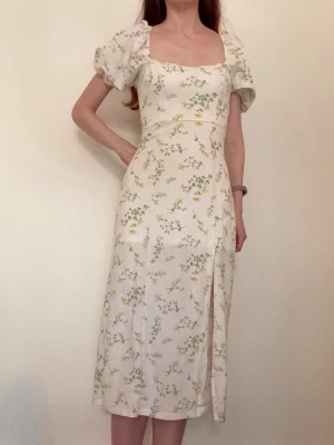 Vit blommig klänning med puffärm - Romantisk vit klänning med grönt och gult blommönster, fyrkantig urringning och puffiga kortärmar. Klänningen har en slits framtill och är figurnära upptill med lösare kjol. Perfekt för sommarens alla tillfällen. Ej stretch. Underkjol som gör att den inte blir genomskinlig. Material viskos och polyamid.