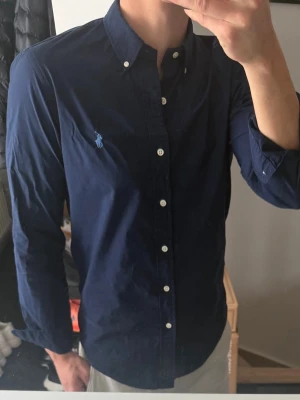 Mörkblå skjorta från Ralph Lauren - Snygg mörkblå skjorta från Ralph Lauren med klassisk button-down krage och vita knappar. Skjortan har lång ärm och ikonisk broderad logga på bröstet. Perfekt för en stilren look och passar till många olika outfits.