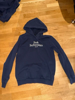 Mörkblå hoodie från Peak Performance - Snygg mörkblå hoodie från Peak Performance med vit broderad logga på bröstet. Klassisk modell med huva och känguruficka framtill. Perfekt för en avslappnad och sportig stil. Mjuk bomullsblandning som känns skön mot huden.