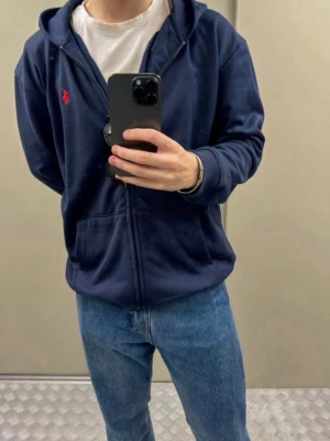 Mörkblå Zip-hoodie - Snygg mörkblå hoodie med dragkedja. Klassisk design med huva, kängurufickor och röd broderad logga på bröstet. Tillverkad i mjuk bomullsblandning som är skön att ha på sig. Perfekt till jeans eller sweatpants.