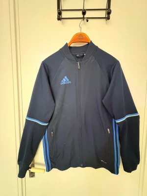 Mörkblå träningsjacka från Adidas - Snygg mörkblå träningsjacka från Adidas med ljusblå detaljer och dragkedja framtill. Jackan har två fickor med dragkedja och klassiska Adidas-loggan på bröstet. Tillverkad i ett lätt och funktionellt material, perfekt för träning eller sport.