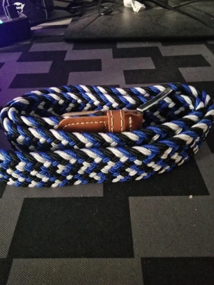 Flätat blått och svart bälte - Säljer ett snyggt flätat bälte i blått, svart och vitt med bruna skinn-detaljer vid spännet. Bältet har ett klassiskt spänne i metall och passar perfekt till jeans eller chinos. Ger en cool och sportig touch till din outfit.