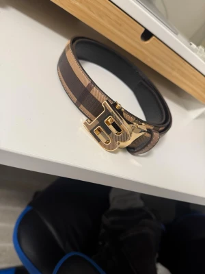 Brunt Burberry bälte med guldspänne - Snyggt bälte från Burberry med klassiskt brunt och beige rutigt mönster och ett stort guldigt B-spänne. Bältet har detaljer i skinn och ger en lyxig känsla till outfiten. Perfekt accessoar för att lyfta din stil.