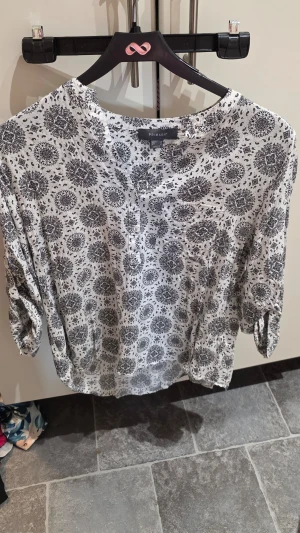 Vit mönstrad blus från Primark - Vit blus från Primark med svart paisleymönster och diskreta knappar framtill. Blusen har trekvartsärm med knappdetalj och är tillverkad i ett lätt och luftigt tyg, perfekt för en avslappnad och trendig look.