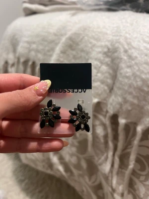 Svarta blomformade örhängen - Snygga örhängen från Accessorize i form av blommor med svarta och silvriga stenar. De har en cool, geometrisk design och är perfekta för att ge din look lite edge. Materialet är metall och plaststenar.