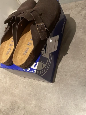Birkenstock Boston mörkbruna tofflor - Säljer ett par Birkenstock Boston tofflor i mörkbrunt mockaläder. Klassisk slip-in modell med justerbart spänne i metall och bekväm korksula. Perfekta för dig som gillar stilrena och tidlösa skor med skön passform. Passar även 44!